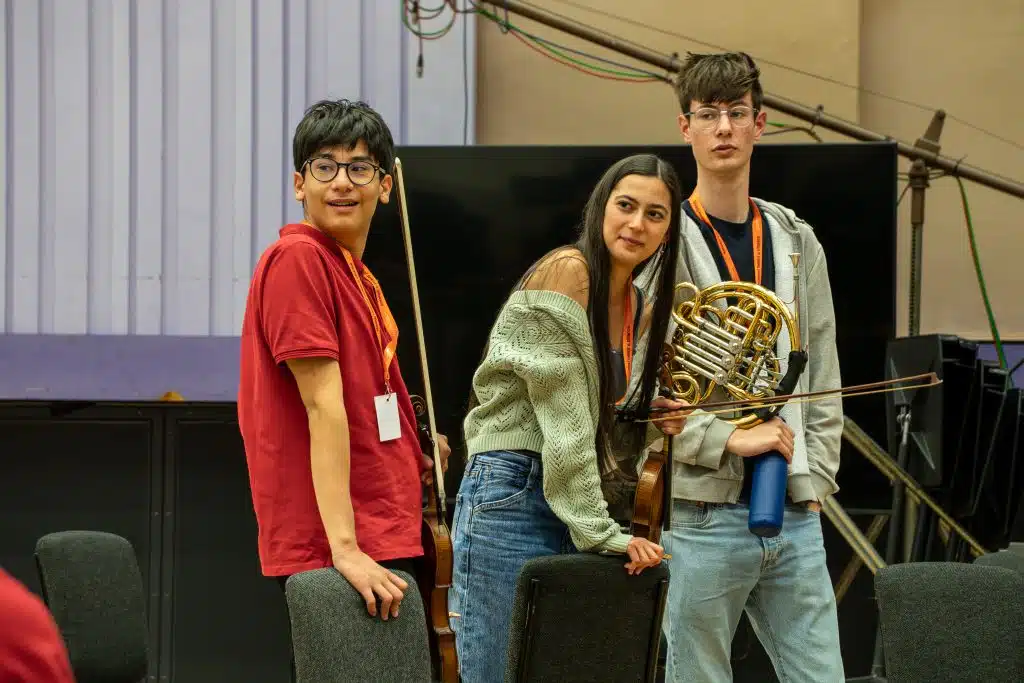 Three young people standing and leaning on chairs looking to their right at Young Sounds' Exchange event in BBC Media City.