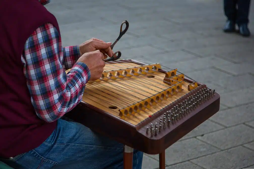 A man in a red, white and blue checkered shirt and red gilet, sat down playing a cimbalom, an uncommon instrument, on the street.
